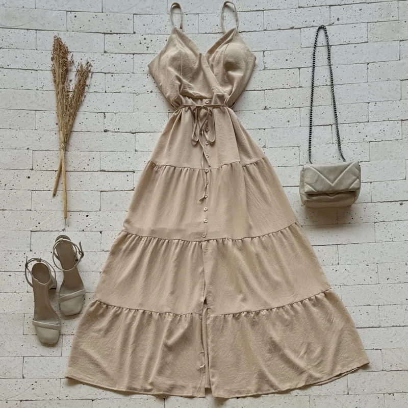Vestido longo caqui com babados, botões frontais, alças finas, amarração na cintura, sandálias e bolsa ao lado sobre piso de pedra.